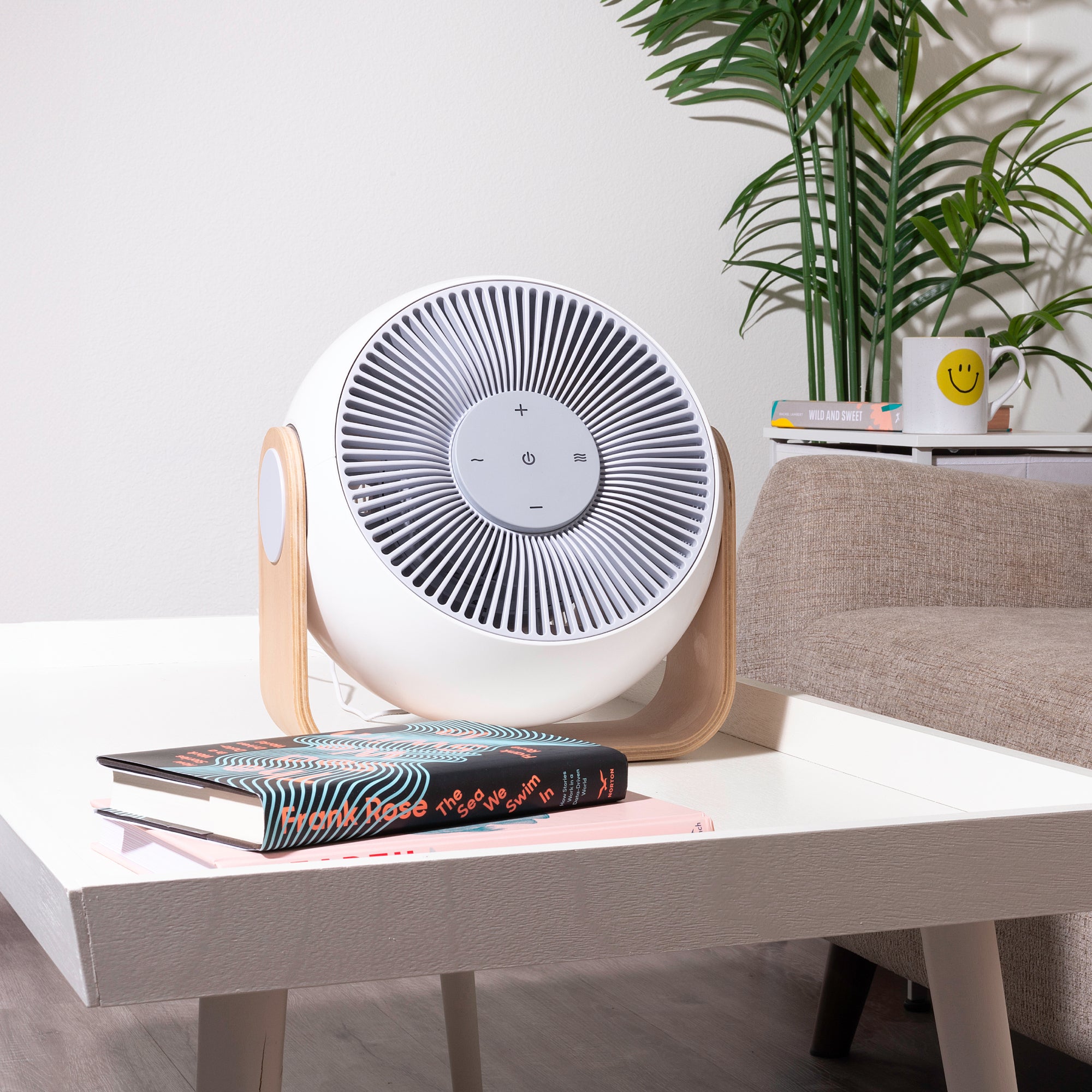 A modern tabletop fan on a white coffee table with books and plants in the background.