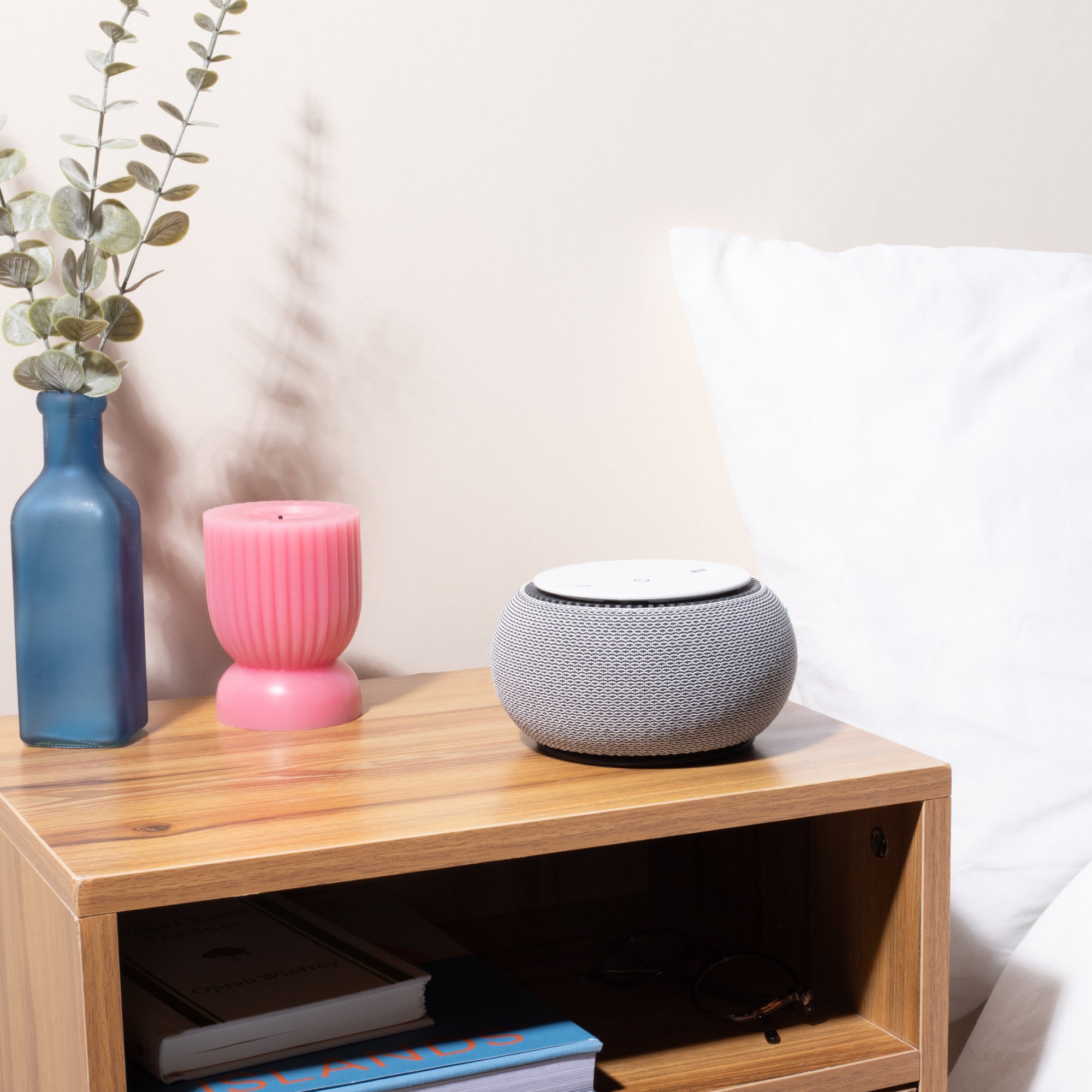 Smart speaker on a wooden nightstand with a pink candle, blue vase, and books.